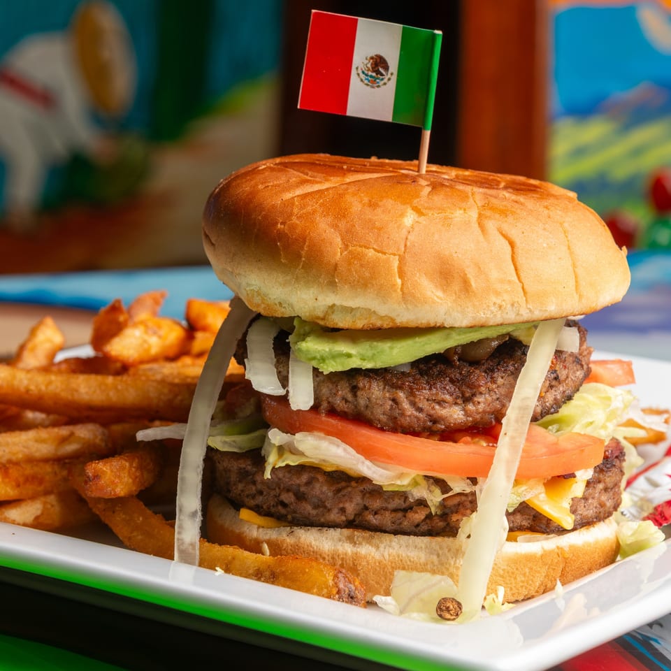 A double-patty burger with avocado, tomato, onion, and a Mexican flag pick, served with fries.
