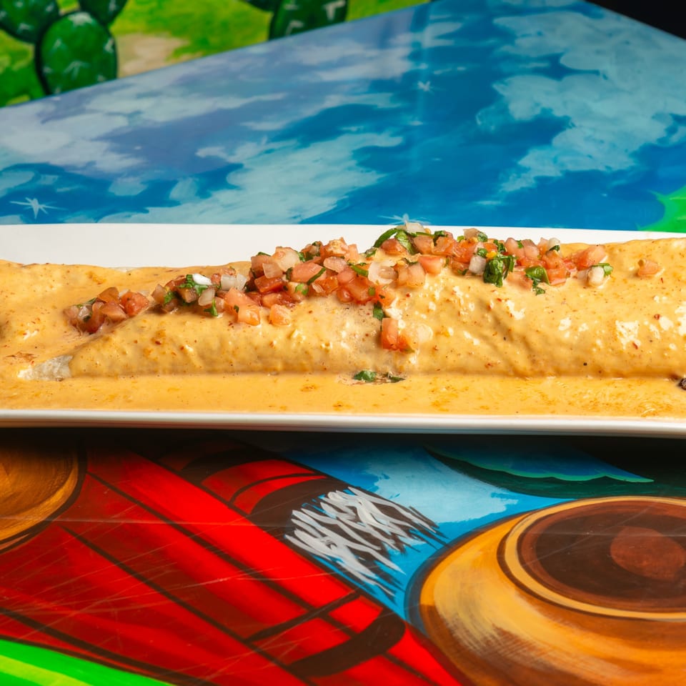 A Magueys Mexican Restaurant burrito covered in queso and pico de gallo on a colorful table.