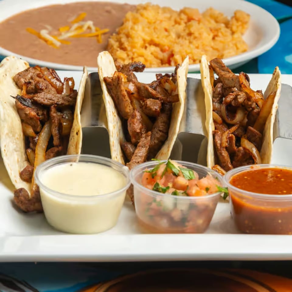 Three steak and onion tacos with sides of rice, beans, salsa, and queso on a white plate.